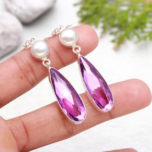 May include: A pair of dangle earrings featuring a pear-shaped purple gemstone and a white pearl at the top of each earring.