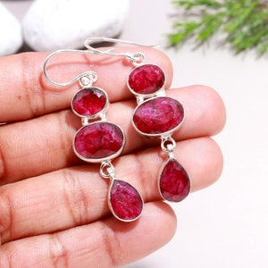 May include: A pair of silver dangle earrings with three red ruby gemstones on each earring. The earrings have a teardrop-shaped ruby gemstone dangling from the bottom.