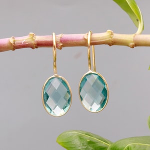 May include: A pair of gold-colored drop earrings with oval, faceted, light blue gemstones. The earrings are suspended from a thin gold wire and are displayed against a blurred background with a plant stem.
