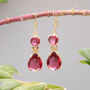 Puede incluir: Pendientes colgantes dorados con piedras preciosas de color rojo-rosa. Cada pendiente presenta una piedra redonda en la parte superior, un elemento de conexión y una gran piedra en forma de lágrima en la parte inferior. Los pendientes cuelgan de un gancho dorado.