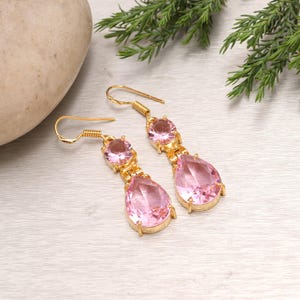 May include: A pair of gold-colored drop earrings with pink gemstones. Each earring features a round gemstone at the top, connected to a larger, teardrop-shaped gemstone below. The earrings are displayed on a light-colored surface with greenery.