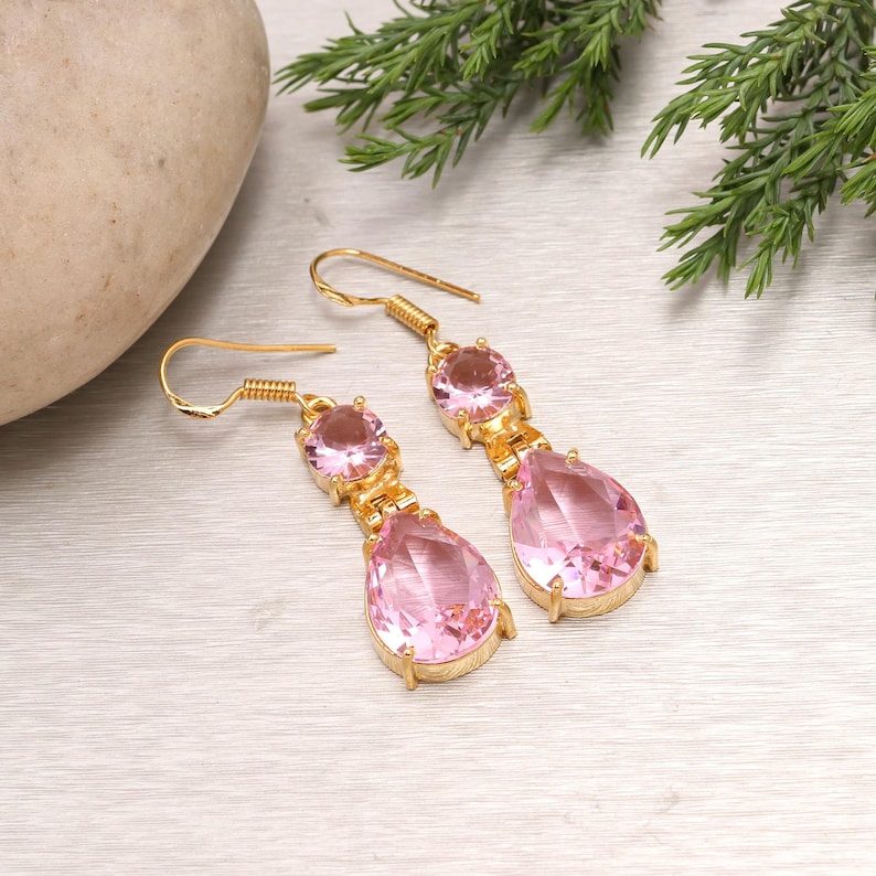 May include: A pair of gold-colored drop earrings with pink gemstones. Each earring features a round gemstone at the top, connected to a larger, teardrop-shaped gemstone below. The earrings are displayed on a light-colored surface with greenery.