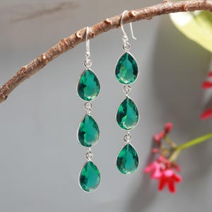 Op de afbeelding: Zilveren oorbellen met drie smaragdgroene, druppelvormige stenen die aan elke haak hangen. De oorbellen hangen aan een natuurlijke tak tegen een zachte grijze achtergrond, met een paar rode bloemen op de achtergrond.