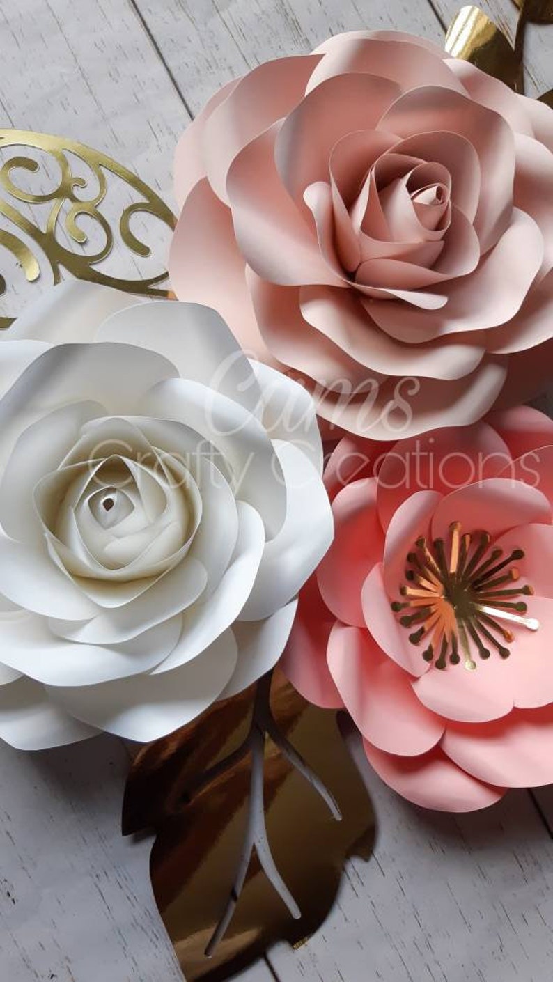 May include: Three large paper flowers in shades of pink and white. The flowers are layered and have a delicate, ruffled texture. The flowers are arranged on a white wooden background.