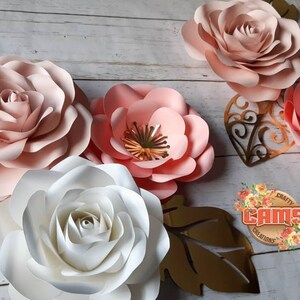 May include: Five paper flowers in shades of pink and white, with gold leaf accents. The flowers are arranged on a white wooden background. The text "CRAFTY CAMS CREATIONS" is visible in the lower right corner.