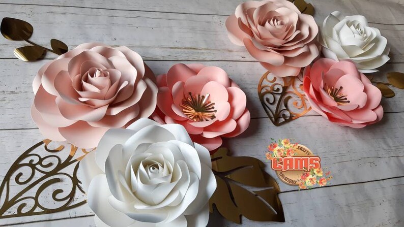 May include: Five paper flowers in shades of pink and white, with gold leaf accents. The flowers are arranged on a white wooden background. The text "CRAFTY CAMS CREATIONS" is visible in the lower right corner.