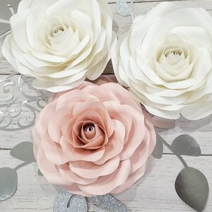 May include: Three paper flowers, one white, one pink, and one ivory, with silver glitter leaves on a white wood background.