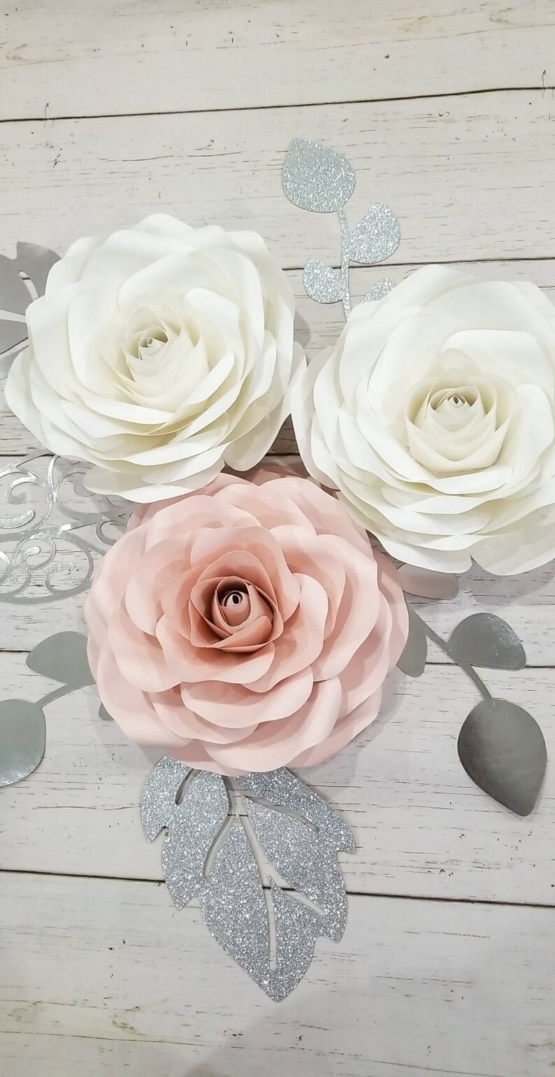 May include: Three paper flowers, one white, one pink, and one ivory, with silver glitter leaves on a white wood background.