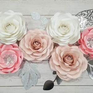 May include: A set of six paper flowers in shades of pink and white, with silver glitter leaves. The flowers are arranged on a white wooden background.