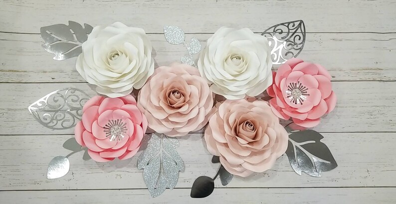 May include: A set of six paper flowers in shades of pink and white, with silver glitter leaves. The flowers are arranged on a white wooden background.