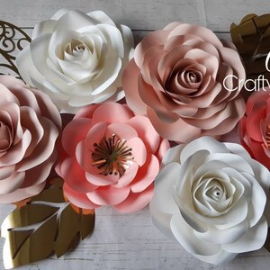 May include: A set of six paper flowers in shades of pink and white. The flowers are arranged on a wooden background with gold leaf accents. The text "Cams Crafty Creations" is visible in the upper right corner of the image.