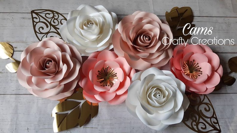 May include: A set of six paper flowers in shades of pink and white. The flowers are arranged on a wooden background with gold leaf accents. The text "Cams Crafty Creations" is visible in the upper right corner of the image.