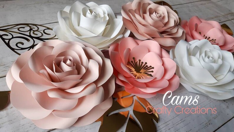 May include: Five large paper flowers in shades of pink and white. The flowers are arranged in a cluster on a wooden surface. The flowers are made of multiple layers of paper and have a delicate, ruffled appearance. The text "Cams Crafty Creations" is visible in the lower right corner of the image.