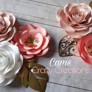 May include: Five paper flowers in shades of pink and white, with gold accents, arranged on a wooden surface. The flowers are layered and have a delicate, romantic feel. The text "Cams Crafty Creations" is visible in the bottom center of the image.