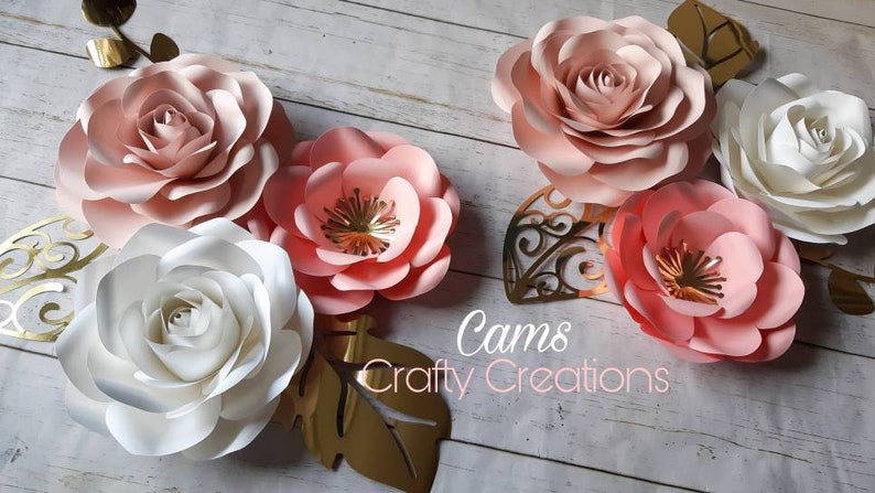 May include: Five paper flowers in shades of pink and white, with gold accents, arranged on a wooden surface. The flowers are layered and have a delicate, romantic feel. The text "Cams Crafty Creations" is visible in the bottom center of the image.