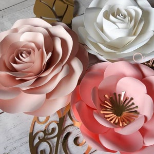 May include: Three paper flowers in shades of pink and white. The flowers are layered and have a delicate, romantic look. The text "Cams Crafty Creations" is visible in the image.