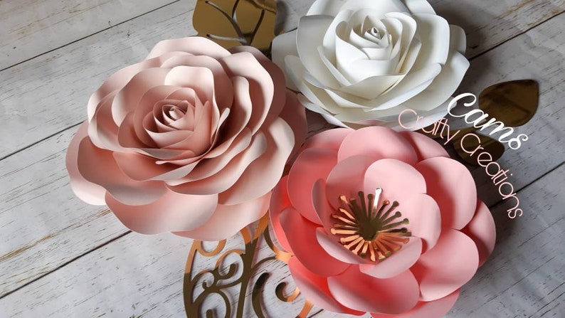 May include: Three paper flowers in shades of pink and white. The flowers are layered and have a delicate, romantic look. The text "Cams Crafty Creations" is visible in the image.