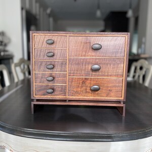 May include: A wooden box with multiple drawers, each with a dark oval knob. The box is made of a dark wood with a light wood inlay. The box is sitting on a dark table.