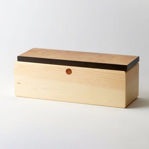 May include: A wooden box with a light wood body and a dark wood top. The box has a small, round, red accent on the front.