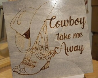 May include: A wooden sign with a cowboy boot and hat design, featuring the text "Cowboy take me Away" in a decorative font. The artwork is burned into the wood, creating a rustic, country-western aesthetic. The sign is displayed on a small white easel.