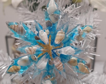 Puede incluir: Un adorno de copo de nieve azul con conchas marinas y una estrella de mar incrustadas en su interior. El adorno está colgado de un árbol de Navidad artificial blanco.