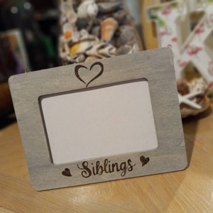 May include: A gray wooden picture frame with a rectangular opening. The frame has a heart carved at the top and the word "Siblings" with two hearts carved at the bottom. The frame is sitting on a wooden surface.