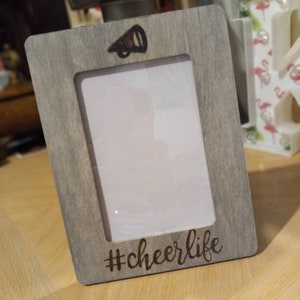 May include: A gray wooden picture frame with a white mat and a black megaphone graphic. The frame has the text "#cheerlife" engraved on the bottom.