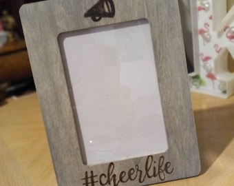 May include: A gray wooden picture frame with a rectangular opening. The frame has a megaphone graphic at the top and the text "#cheerlife" at the bottom. The frame is leaning against a light-colored surface.