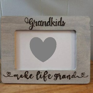 May include: A rectangular gray wooden picture frame with the words "Grandkids" and "make life grand" in black script. The frame has a heart-shaped cutout in the center for a photo. A heartwarming gift for grandparents.