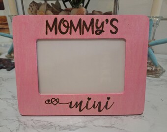 May include: Pink wooden picture frame with the words "Mommy's mini" etched in dark brown. The frame has a rectangular opening and is decorated with a heart and swirl design.