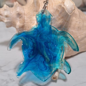 May include: A clear resin turtle-shaped keychain with swirling blue and white patterns. The keychain has a silver metal ring and chain. The background features a seashell.