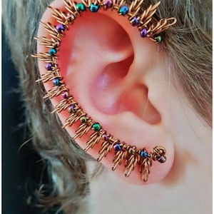 May include: A copper ear cuff with iridescent beads in shades of purple, green, and blue. The cuff is shaped like a crescent moon and has a delicate, spiky design.