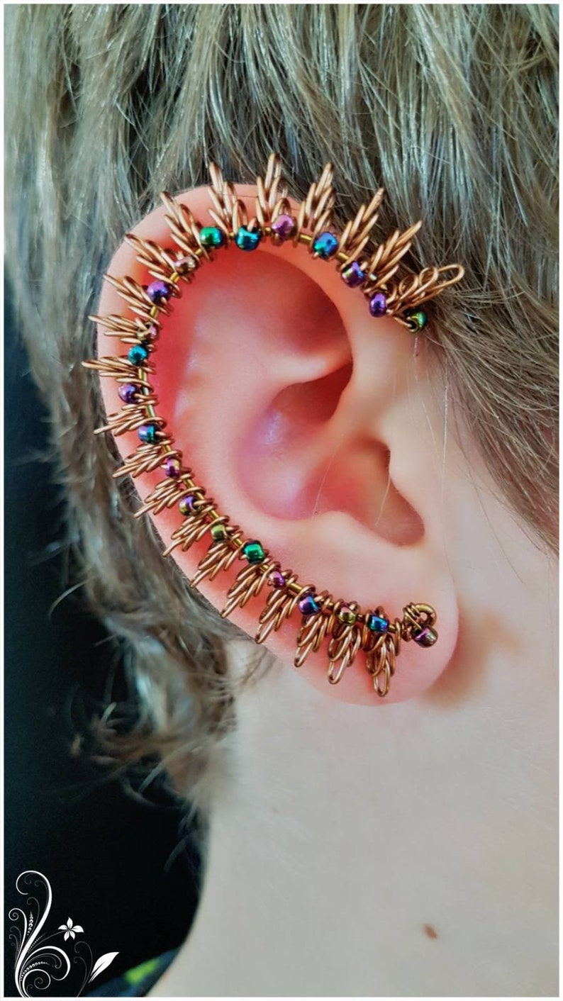 May include: A copper ear cuff with iridescent beads in shades of purple, green, and blue. The cuff is shaped like a crescent moon and has a delicate, spiky design.