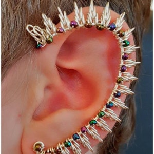 May include: A silver ear cuff with a row of iridescent beads in shades of green, purple, and brown. The cuff is shaped like a spike and wraps around the upper ear.