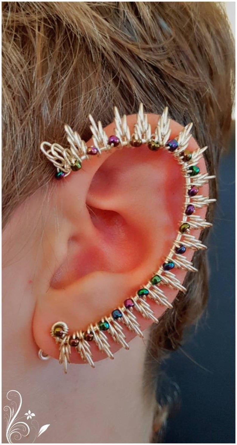 May include: A silver ear cuff with a row of iridescent beads in shades of green, purple, and brown. The cuff is shaped like a spike and wraps around the upper ear.