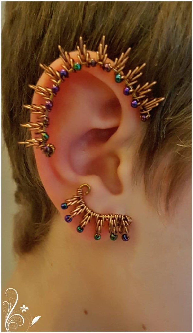 May include: A pair of ear cuffs made of copper wire with small beads. The ear cuffs are shaped like a crescent moon and have a spiky design. The beads are iridescent and come in shades of purple, green, and blue.