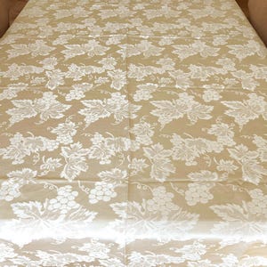 May include: A rectangular tablecloth featuring a tan base color with an all-over white woven pattern of grape clusters and vine leaves. The fabric displays a repeating botanical motif across the entire surface, creating a textured appearance.