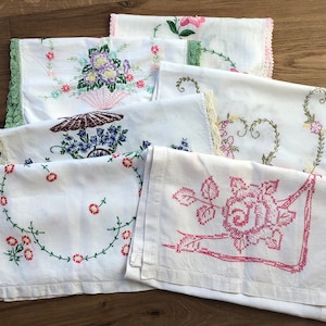 May include: A collection of six white rectangular linens featuring various colorful floral embroidery designs, including roses and baskets, with decorative crocheted or lace edges in shades of green, pink, and cream, arranged in a layered stack.