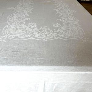 May include: A rectangular white tablecloth featuring a woven damask pattern. The fabric displays a prominent border design composed of intricate, swirling floral and scrollwork motifs that frame the central area of the textile.