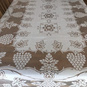 May include: A white and brown lace tablecloth with a grape and leaf design. The tablecloth has a detailed pattern with grapes, leaves, and floral elements. The edges are finished with a delicate lace trim.