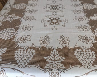 May include: A white and brown lace tablecloth with a grape and leaf design. The tablecloth has a detailed pattern with grapes, leaves, and floral elements. The edges are finished with a delicate lace trim.