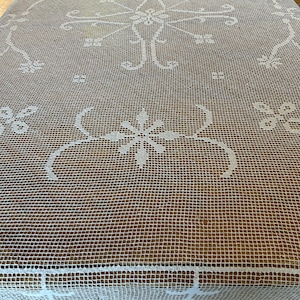 May include: A white, open-weave tablecloth with a detailed floral and scrollwork design. The tablecloth is rectangular and covers a wooden table. The intricate pattern is visible across the entire surface, creating a delicate and elegant aesthetic.