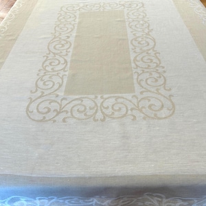 May include: A rectangular tablecloth featuring a light beige center panel surrounded by a wide white border. The fabric displays a repeating scrollwork pattern in a contrasting beige tone that frames the central rectangle and edges.