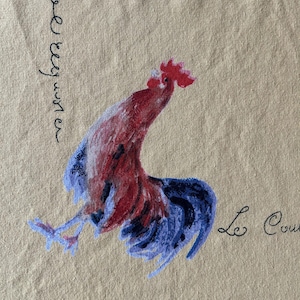 May include: A beige fabric with a hand-painted rooster design. The rooster is depicted in shades of red and blue, with the words "Le Coq" and other text in black script. The artwork has a rustic, country aesthetic.