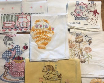 May include: A collection of white and yellow fabric towels featuring various embroidered designs including a dog in a tub, a scarecrow, a basket of flowers, a person with apples, a dog with a tea set labeled CHINA, and wine glasses.