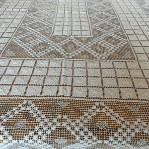 May include: Cream-colored, open-weave tablecloth with intricate geometric and floral patterns. The design features a central rectangular motif, surrounded by diamond and square patterns, and a scalloped edge. The tablecloth is made of a delicate, possibly crocheted or lace material.