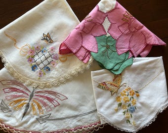 May include: A collection of three white fabric napkins featuring intricate embroidery. One napkin displays a butterfly and floral grid, another shows a large stylized butterfly, and the third features a butterfly with yellow and blue flowers.