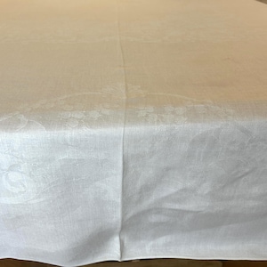 May include: A white rectangular tablecloth featuring a subtle, tonal damask pattern with woven floral and scroll motifs. The fabric has a smooth, matte finish with a visible center seam running vertically down the length of the textile.