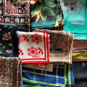May include: A collection of folded square silk scarves featuring various patterns including floral, geometric, and indigenous-inspired designs. The scarves display diverse colors such as black, red, white, green, blue, and brown, with visible brand labels.
