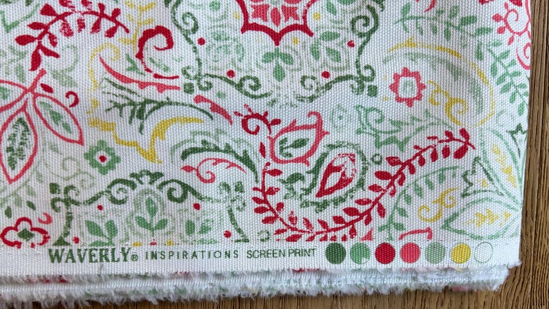 May include: A white fabric featuring a repeating screen-printed pattern of red, sage green, and yellow paisley and floral motifs. The bottom edge displays the text WAVERLY INSPIRATIONS SCREEN PRINT followed by a row of colored circular swatches.